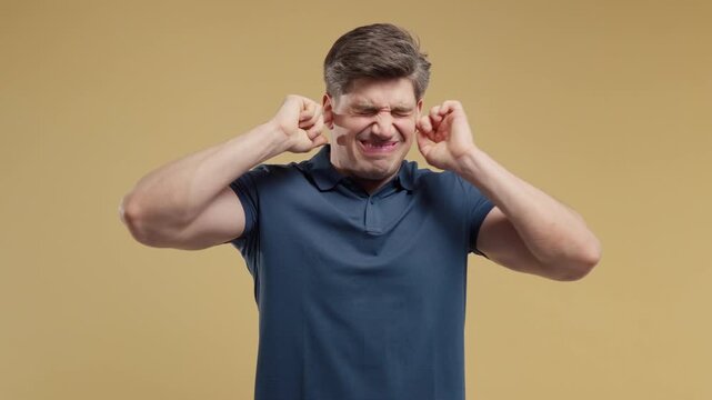 Frustrated young man covering ears to block unwanted noise. Stress, overload, conflict, distraction and mental pressure. Studio portrait on a yellow background