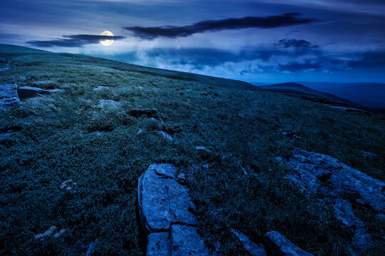 carpathian mountain landscape on summer night. fantasy view of a grassy alpine meadow. full moon over rock and yellow herbs on the hill. mysterious mountain weather with cloudy sky