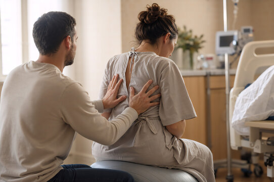 Man doing back massage for his pregnant wife sitting on the birth ball. Partner assisted birth support in hospital room. Couple during labor and childbirth preparation. 
