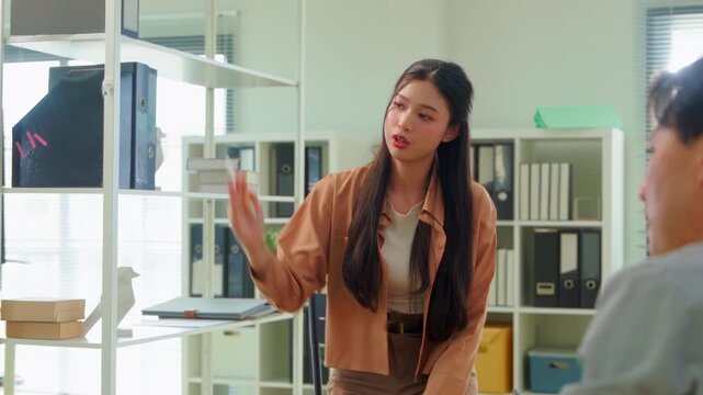 A businessman presents and points to charts pinned on the wall while two female colleagues listen attentively, one typing notes on a laptop, during a team meeting in a bright modern office.