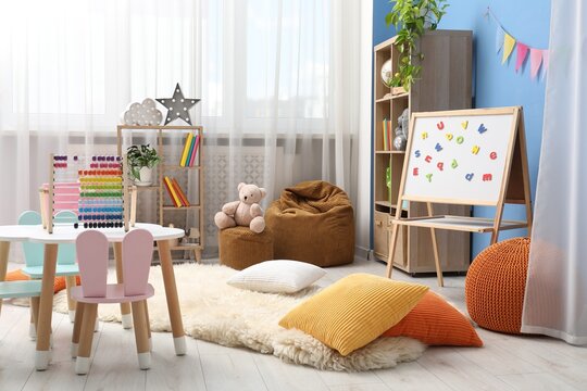 Small table with chairs, shelving units, colorful pillows, poufs and magnetic board in stylish child's room. Interior design