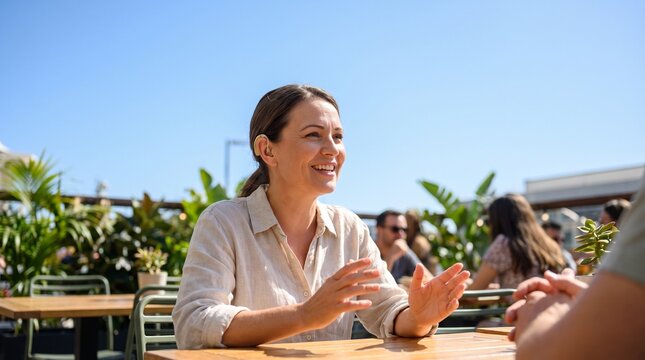 Happy woman wearing hearing aid talking with friend at outdoor cafe. Healthcare technology for deaf person.