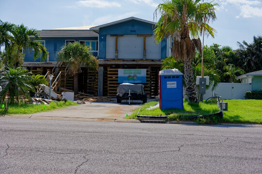 Elevated coastal house on stilts undergoing renovation, with construction debris, a covered boat and a portable toilet by the driveway, framed by palm trees in a sunny suburban setting.
