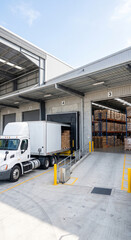 Naklejka premium White delivery truck backed into a warehouse loading bay beside dock doors and pallet stacks in an industrial yard in an industrial logistics warehouse
