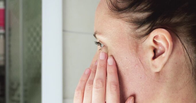 Woman examines facial skin in bathroom mirror while gently touching her face. Close-up captures daily skincare routine, healthy skin assessment, natural beauty and personal self-care.