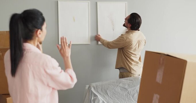 Couple boxes decorating apartment, man hanging pictures. They align framed prints on a light wall amid unpacking, signaling a fresh start at home. Teamwork in home moving and decorating