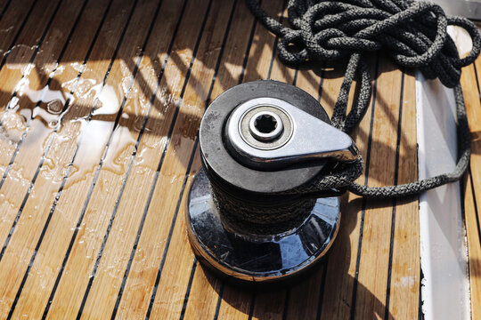 Sailboat winch holds braided rope. Wet wooden deck forms background. Black line wraps metal drum. Marine hardware rests under sunlight. Water drops remain between boards. Deck gear stays ready.