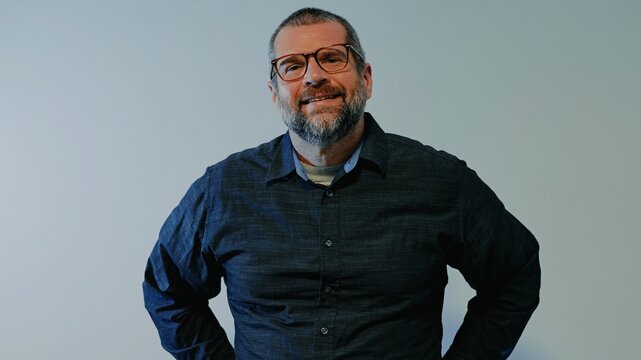 Confident middle-aged man with beard wearing a dark button-down shirt posing against a neutral backdrop, representing modern business leadership
