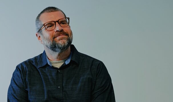 Confident middle-aged man with beard wearing a dark button-down shirt posing against a neutral backdrop, representing modern business leadership