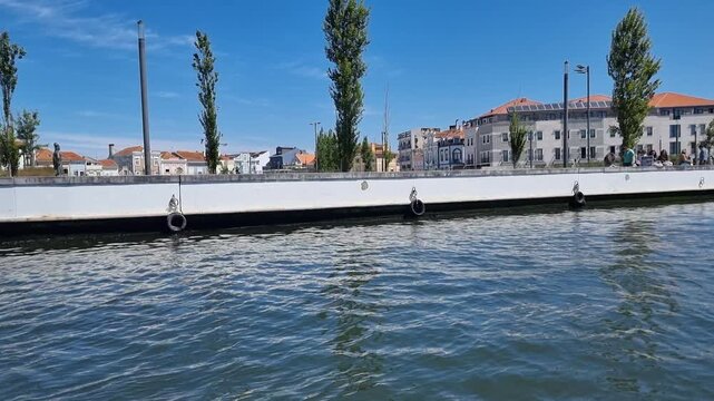 POV shot from a boat sailing along the central canal in Aveiro, Portugal, showing the waterfront promenade, city architecture, and trees under a clear blue sky on a bright sunny day.