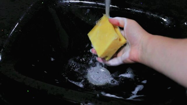 Hand cleaning a black kitchen sink with a soapy cleaning sponge, white foam covering the sink surface during a thorough home cleaning and disinfection routine today