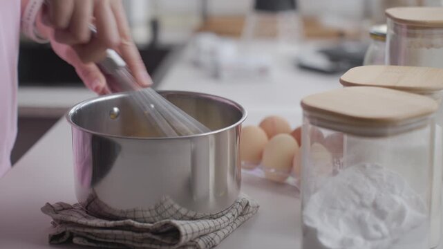 Woman whisks ingredients in metal saucepan on kitchen counter, with eggs and ingredient jars nearby. Concept of home baking, cake recipe and food preparation