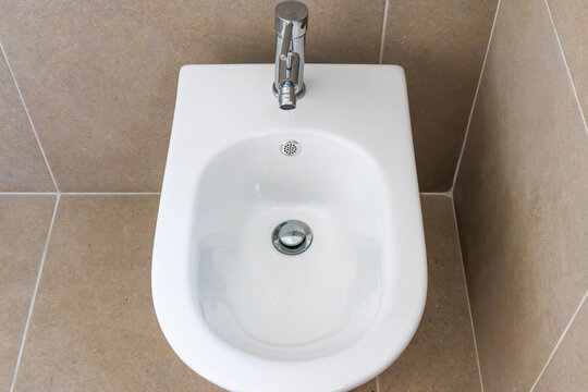 Modern white bidet with chrome faucet and drain in a tiled bathroom
