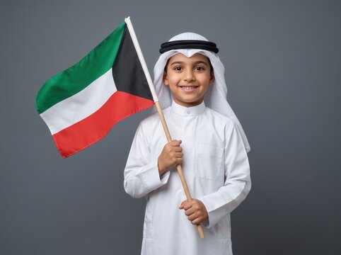 Young boy wearing traditional white thobe and ghutra holding a kuwaiti flag against a solid grey background while smiling at the camera with a cheerful expression and upright posture