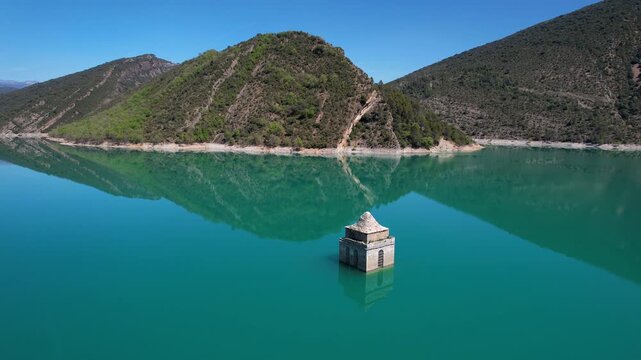 Orbital aerial shot of Asunci&oacute;n de Mediano Church covered with water of Mediano Reservoir and snow-covered Pyrenees on sunny spring day. Huesca, Spain.