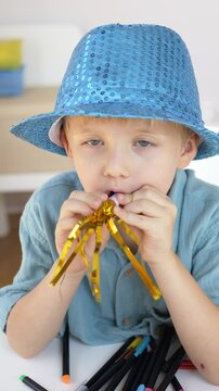 Boy in blue hat blows golden tinsel noisemaker with eyes closed in immersed vertical birthday portrait. Gold festive celebration and joyful birthday party concept. 