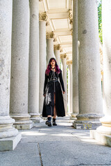 Naklejka premium Young woman in gothic attire walking confidently through a colonnade, dressed for a carnival, masquerade or Halloween celebration, blending alternative fashion with historic architecture