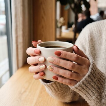 Close-up of woman's hands with elegant matte terracotta and beige manicure holding a ceramic mug of hot coffee in a cozy cafe setting, wearing a warm knit sweater