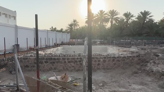 Fenced archaeological excavation site near historic ruins and palm trees at sunset.