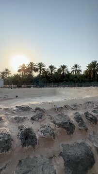Historic stone pool ruins surrounded by lush palm trees under a bright sunset sky.