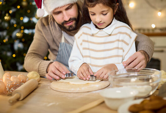 Father and daughter bake Christmas cookies at home. In a cozy kitchen with tree lights, man and child make dough and cut shapes, sharing joy and baking together. Warm family winter holidays.