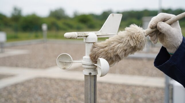 Person wearing gloves using a fluffy duster to clean a white weather station anemometer outdoors