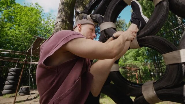 Caucasian young adult man stretching leg on tire obstacle wall at outdoor gym. Footage is perfect for promoting outdoor fitness programs, athletic training, and active lifestyle brands