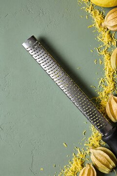 Overhead flat lay of a metal microplane grater on a green textured background, surrounded by scattered yellow lemon zest and dry physalis husks.