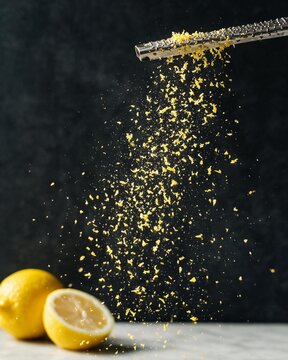 A dynamic close-up of yellow lemon zest falling like a shower of gold dust from a microplane grater, with whole and halved lemons in the foreground.
