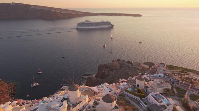 Drone aerial footage of traditional windmills and white houses in Oia at sunset with cruise ship sailing caldera sea Santorini Greece