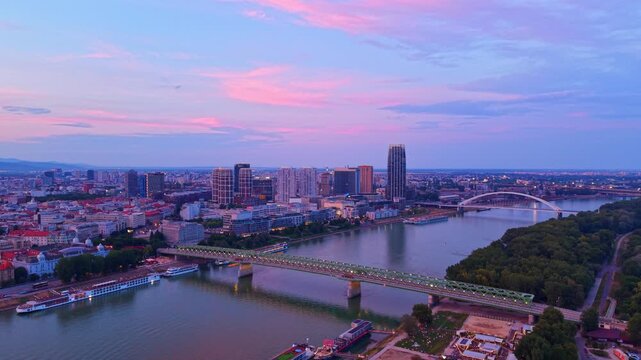 View of city skyline and river at dusk. City buildings and a river are shown as the sun sets in the evening sky. Lights start to glow in the city.