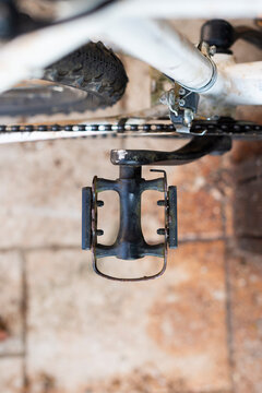 A vertical top-down view shows a used black bicycle pedal attached to a crank arm. Parts of the white frame, chain, and tire are visible over a paved surface.