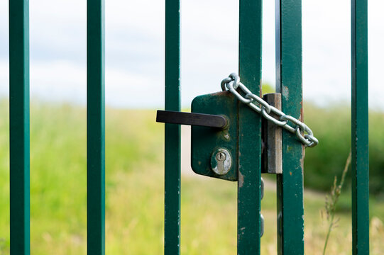 A close-up shot shows a green metal gate secured with a thick chain and built-in lock. The blurred background features a green grassy field under an overcast sky.