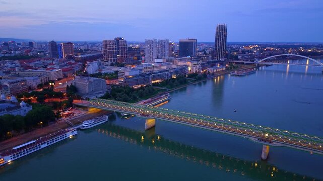 City view with bridge and river at dusk. A river flows beside a city skyline as buildings light up in the evening. People enjoy the view from the bridge.
