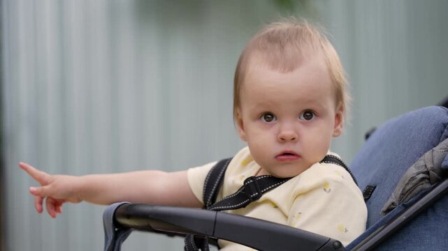 Baby seated in stroller gazes curiously around outdoor setting. Starts looking sideways then turns head toward camera with surprised expression before looking away again