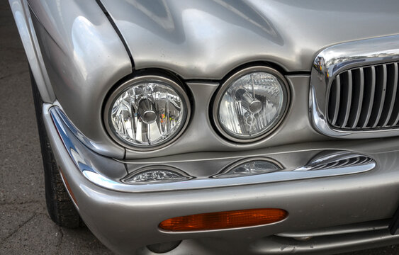 Close up of a silver Jaguar front end. It highlights the iconic dual round headlights, a contoured hood, an amber turn signal, and a curved chrome grille