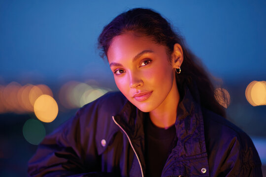 Young woman in a dark bomber jacket illuminated by purple and orange neon light against a blue night city sky