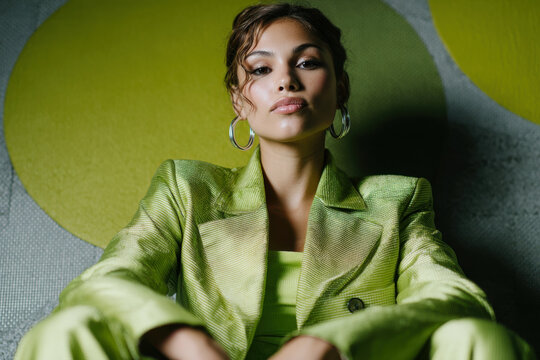 Woman sitting on floor wearing oversized lime green blazer and hoop earrings against green abstract wall