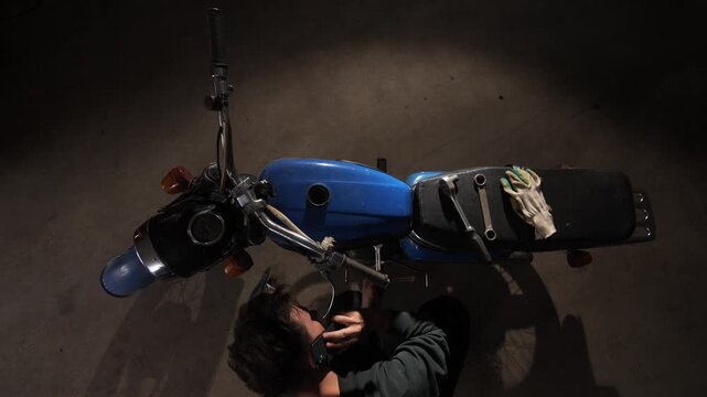 Man in garage holding smartphone to ear and talking while working on engine of a classic blue motorcycle, surrounded by tools and equipment