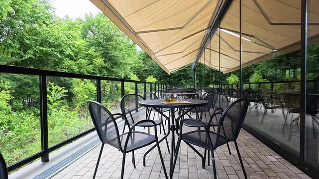 Outdoor patio with tables and chairs under a beige awning. Green trees surround the glass-walled balcony. Black metal chairs sit on a tiled floor. No people are visible in the scene
