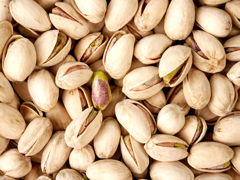 A close-up, overhead view of a large pile of roasted pistachios. One pistachio is split open, revealing its green nut.
