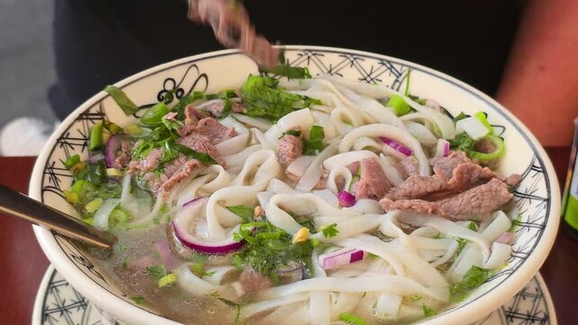 Chopsticks lift a piece of beef from a bowl of pho noodle soup, fresh herbs and onions visible.