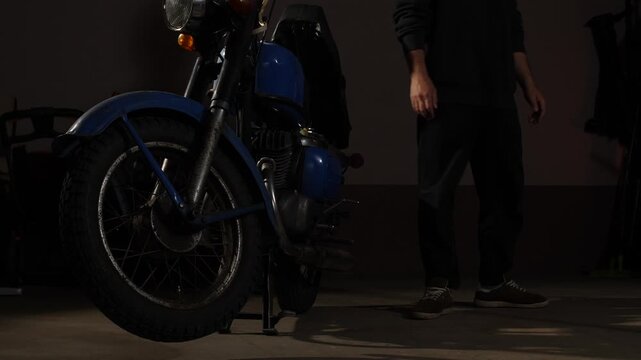 Man removing work gloves after completing maintenance on a classic blue motorcycle, highlighting dedication and the joy of a mechanical hobby in a dimly lit workshop