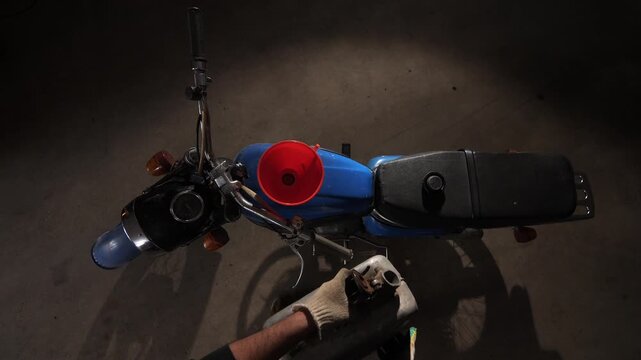 Blue vintage 1970 motorcycle preparing for fuel refill in a dimly lit garage, showing a hand wearing a glove holding a jerry can, funnel placed in the tank opening