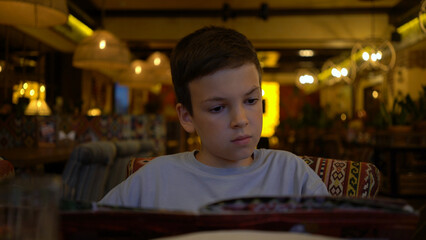 Young boy sits in cozy restaurant, carefully reading menu and choosing what to eat, focused expression under warm ambient lighting. © MrP