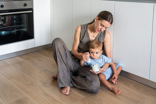 Mother feeding toddler son yogurt on kitchen floor at home