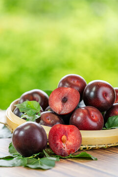 Red Plums on the wooden table over blurred greenery background, Fresh Juicy plums in wooden basket on wooden Background.