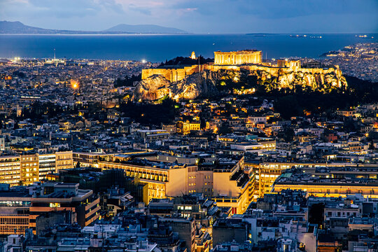 Stunning night view of the Acropolis and the Parthenon brilliantly lit up, overlooking the densely built city of Athens city, Greece. Sea and islands visible in distance. Viewpoint on Philopappos Hill