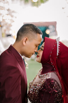 Indonesian Muslim Wedding Couple in Maroon Attire Forehead Touch Intimate Portrait with Hijab Bride and Groom in Suit