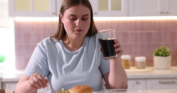Overweight woman eating French fries and drinking cola at light marble table in kitchen. Camera moving forward